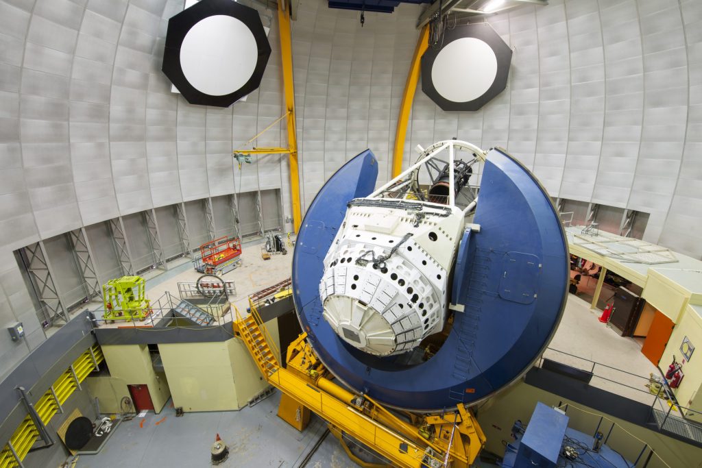 A large blue disc with the top portion open has a has a big white instrument sitting at its center. This instrument sits atop a yellow stand. In a large room with a flight of stairs, the whole instrument is large, much taller than the flight of stairs and equally wide.