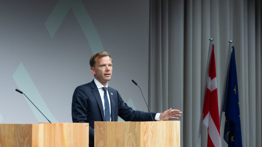 Man in suit at lectern, behind him flags.